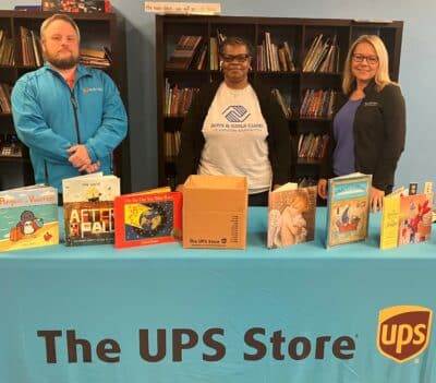 1 man and 2 women are standing behind a table with a table cloth showing The UPS Store logo. On the table, there are books standing up.