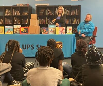 A woman is standing behind a table with a table cloth displaying The UPS Store logo. There are books standing on the table. There is a man seated next to her. She is presenting to youth sitting on the floor looking at her.