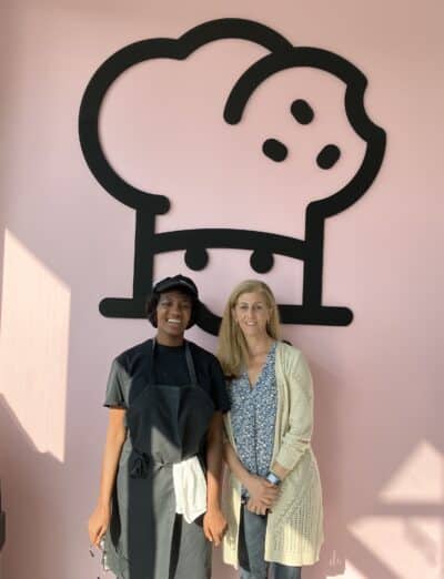 Camara and Laura standing in front of large Crumbl Cookies logo on a pink wall