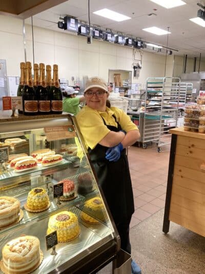 Maggie leaning against the bakery display case with arms folded, wearing an apron, looking at the camera and smiling.