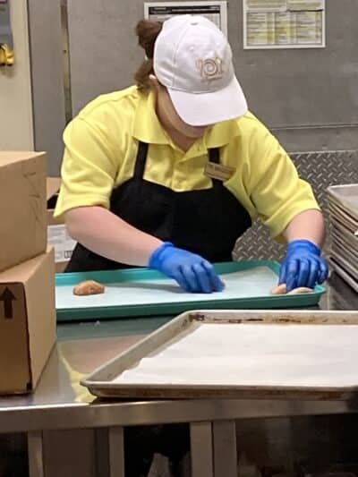 Maggie is wearing a baseball cap and blue gloves, placing cookies on a cookie sheet.