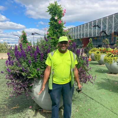 Mikey stands in front of a large plant at BWI. He is wearing a neon green shirt and crew vest.