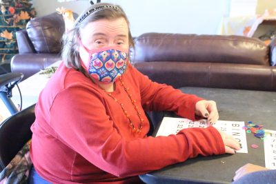 A woman in pink shirt, wearing a mask, headband and necklace looks up at the camera during a Bingo game