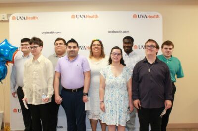 Group of high school students standing in front of a UVA Health background. They are looking at the camera. 
