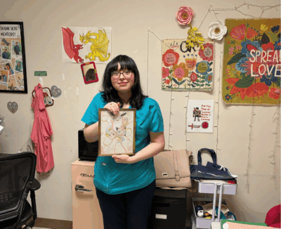 Verali is holding up a piece of artwork, standing in front of a wall of art in an internship classroom.